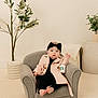 baby, child, armchair, trench_coat, headband, starbucks_cup, indoor, plant, pottery, minimalist, cozy, carpet, footwearless, cute, portrait, furniture, expression, fashion, relaxing, neutral_colors