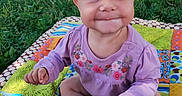 Catherine is registered to the contest to win money with this photo: baby, child, sitting, smiling, happy, portrait, outdoor, grass, blanket, patchwork, colorful, purple_clothing, floral_pattern, blue_eyes, blond_hair, infant, hand, barefoot, parked_car, sunlight