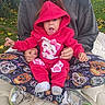 baby, child, infant, pink_hoodie, hooded_sweatshirt, hello_kitty, sitting, adult, parent, hands, surprised, open_mouth, portrait, outdoor, grass, dandelions, fallen_leaves, cushion, patterned_fabric, sneakers