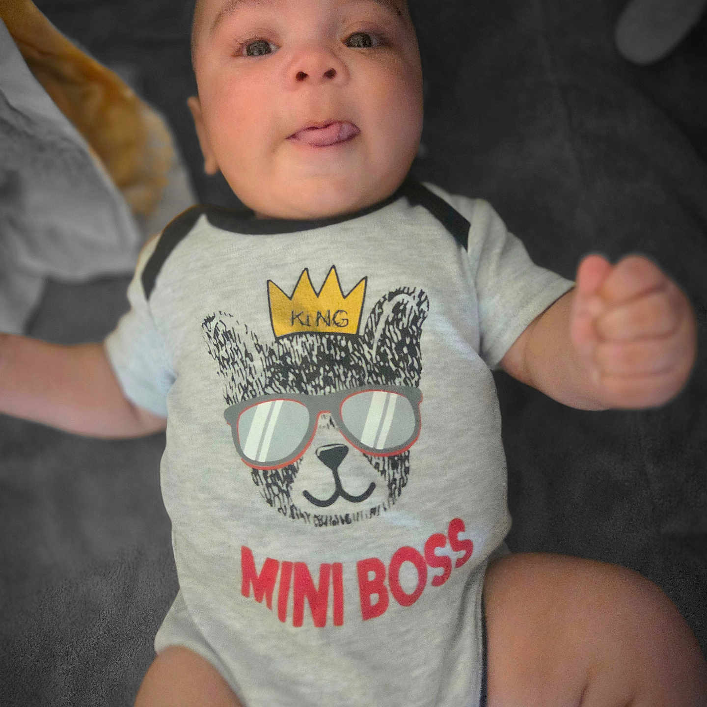 Atrayus is registered to the contest to win money with this photo: apparel, baby, child, clothing, cute, expression, face, fist, graphic_tee, indoors, infant, lying_down, onesie, person, photography, playful, smile, soft_surface, tongue, young