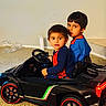 children, boys, siblings, ride_on_car, toy_car, sports_car, indoor, portrait, sitting, driving, steering_wheel, smiling, blue_clothing, red_clothing, carpet, living_room, vehicle, wheels, photograph, playtime