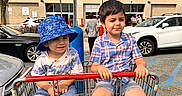 Orhan is registered to the contest to win money with this photo: children, boys, siblings, shopping_cart, outdoor, parking_lot, warehouse_store, costco, blue_hat, bucket_hat, plaid_shirt, shorts, sandals, smiling, portrait, sunlight, cars, tree, shopping, summer