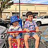 children, boys, siblings, shopping_cart, outdoor, parking_lot, warehouse_store, costco, blue_hat, bucket_hat, plaid_shirt, shorts, sandals, smiling, portrait, sunlight, cars, tree, shopping, summer
