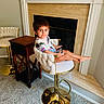 child, toddler, portrait, sitting, smiling, indoors, fireplace, chair, furniture, phone, smartphone, living_room, home, carpet, gold_trim, cream_decor, table, barstool, casual_clothing, barefoot