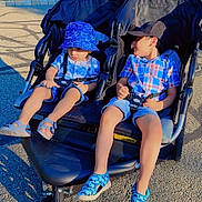Orhan joined the competition — help win amazing prizes! children, toddler, siblings, stroller, outdoor, sunny, driveway, yard, trees, blue_clothing, sun_hat, baseball_cap, plaid_shirt, shorts, sandals, harness, seated, sibling_bond, water_bottle, family