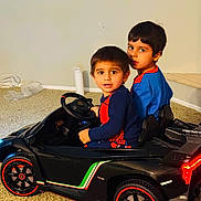 Osman is registered to the contest to win money with this photo: children, boys, brothers, toy_car, ride_on_car, sports_car, indoor, living_room, carpet, sitting, portrait, faces, smiling, looking_at_camera, dark_hair, blue_clothing, red_clothing, steering_wheel, wheels, toy_vehicle