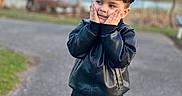 Osman is registered to the contest to win money with this photo: child, boy, portrait, outdoor, park, playground, smiling, face, cheeks, hands, standing, jacket, leather_jacket, jeans, brown_hair, fashion, path, pond, trees, bokeh