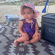 Layla is registered to the contest to win money with this photo: toddler, child, beach, sand, sunhat, swimsuit, mat, cooler, summer, vacation, outdoor, pattern, water, toy_box, sunny, building, relaxation, barefoot, playful, smiling
