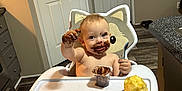 Odin is registered to the contest to win money with this photo: toddler, child, high_chair, chocolate, messy, face_paint, spoon, snack, kitchen, flooring, door, cabinet, countertop, indoor, person, happy, smiling, cute, baby, feeding