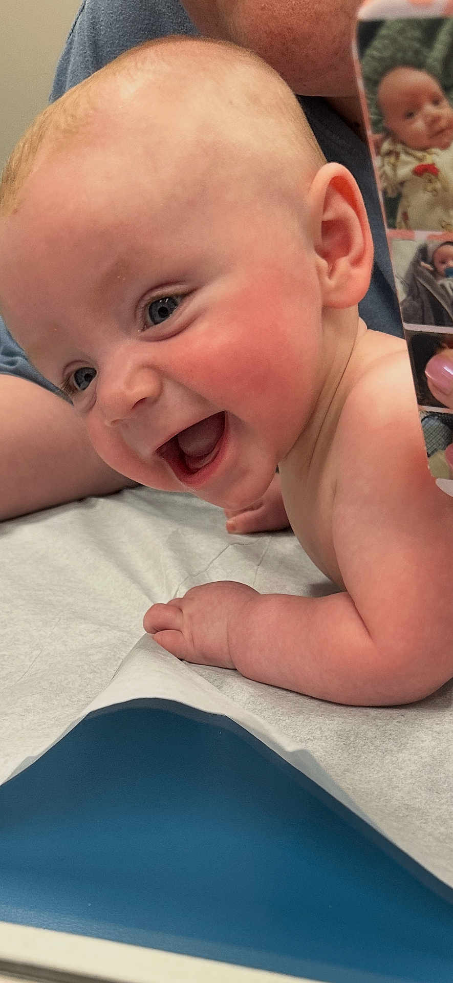 Harrison joined the competition — help win amazing prizes! baby, smiling, child, infant, face, cheeks, skin, arm, hand, medical_table, phone, photo, reflection, adult, background, happy, closeup, expression, person, healthcare