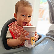 Eyden a rejoint le concours — aidez-le/la à gagner de superbes lots ! baby, child, high_chair, sippy_cup, bib, smiling, happy, indoor, cute, toddler, face, hands, drink, furniture, person, young_child, expression, table, baby_bib, water