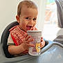 baby, child, high_chair, sippy_cup, bib, smiling, happy, indoor, cute, toddler, face, hands, drink, furniture, person, young_child, expression, table, baby_bib, water