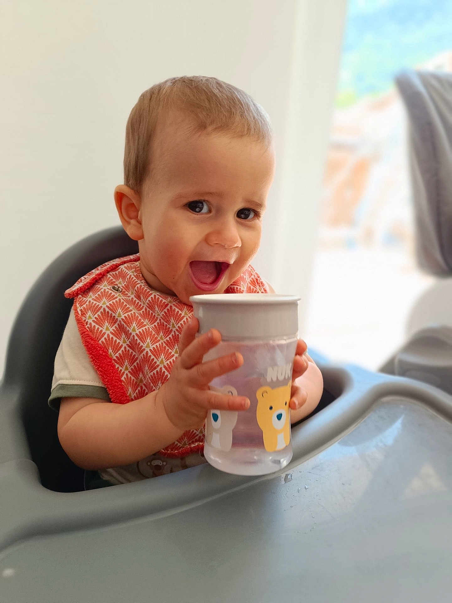 Eyden a rejoint le concours — aidez-le/la à gagner de superbes lots ! baby, child, high_chair, sippy_cup, bib, smiling, happy, indoor, cute, toddler, face, hands, drink, furniture, person, young_child, expression, table, baby_bib, water