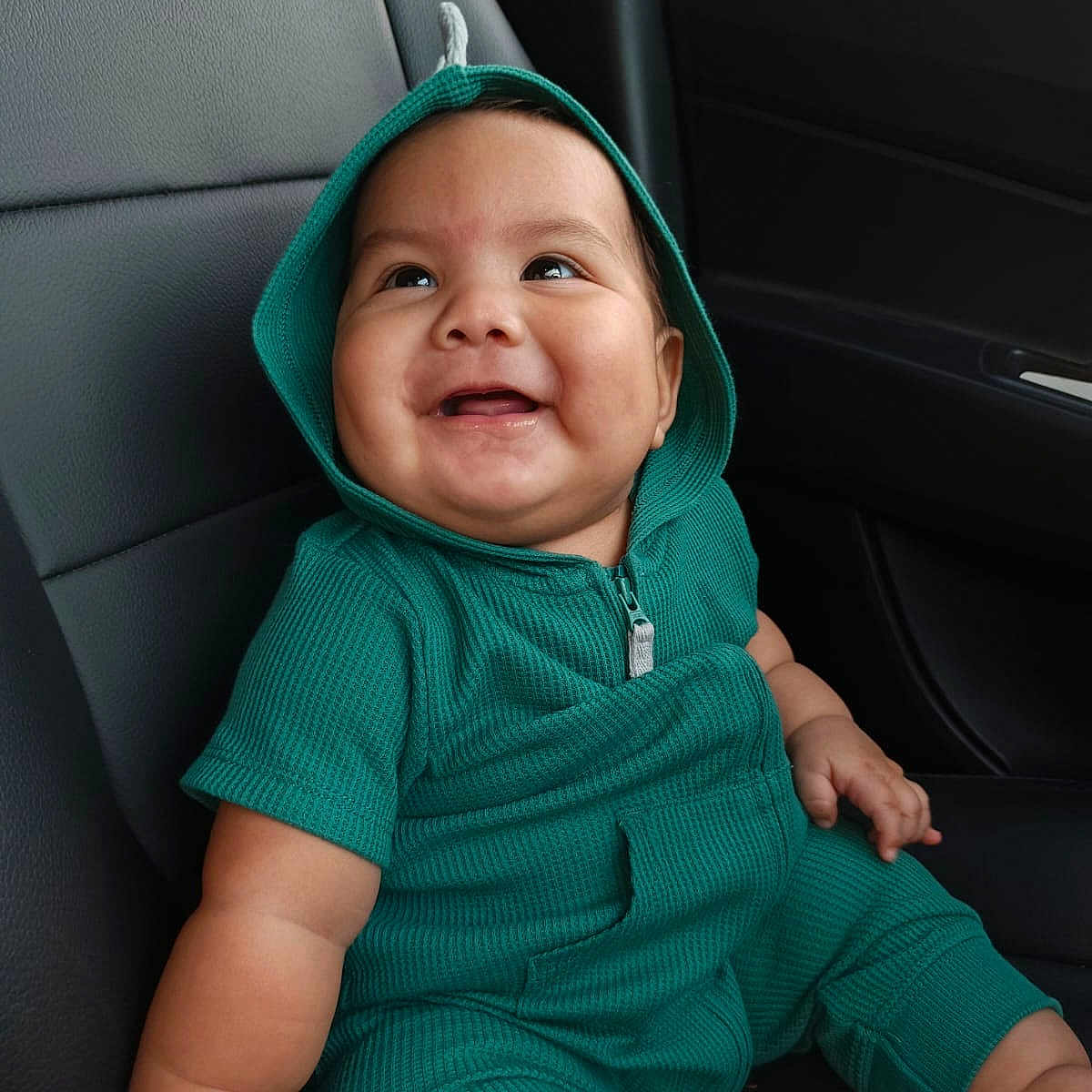 Santiago is registered to the contest to win money with this photo: baby, car_interior, car_seat, child, clothing, cute, expression, green_hoodie, happy, hood, indoors, infant, joy, person, portrait, seat, sitting, skin, smiling, young_child