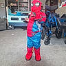 child, costume, spiderman, indoor, carpet, bed, shelf, toy, wheelchair, furniture, living_room, person, playful, pose, resting, home, decor, boots, mask, carpeted_floor