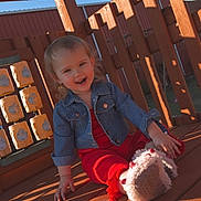 Scarlett is registered to the contest to win money with this photo: toddler, child, smiling, denim_jacket, red_outfit, playground, wooden_deck, toy, steering_wheel, sunlight, shadow, outdoor, happy, person, face, sitting, daylight, cute, play, casual