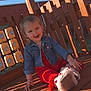toddler, child, smiling, denim_jacket, red_outfit, playground, wooden_deck, toy, steering_wheel, sunlight, shadow, outdoor, happy, person, face, sitting, daylight, cute, play, casual