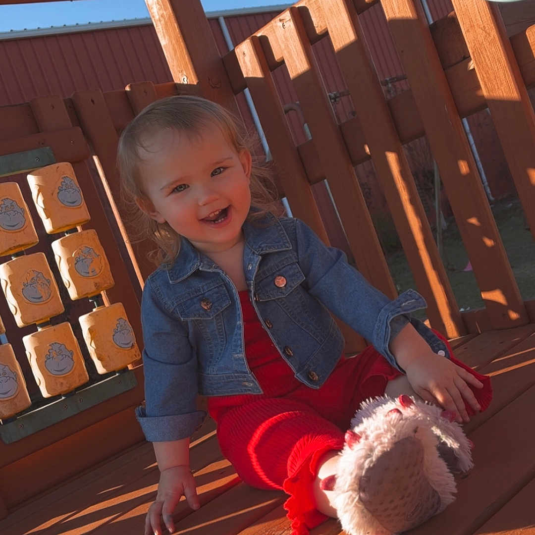 Scarlett is registered to the contest to win money with this photo: casual, child, cute, daylight, denim_jacket, face, happy, outdoor, person, play, playground, red_outfit, shadow, sitting, smiling, steering_wheel, sunlight, toddler, toy, wooden_deck