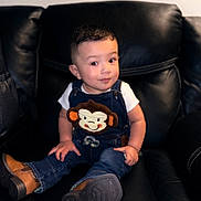 Julian is registered to the contest to win money with this photo: toddler, child, denim_overalls, monkey_patch, boots, leather_couch, smile, curious, indoor, portrait, young_child, fashion, cute, seated, casual_clothing, happy, baby, person, cute_expression, furniture