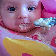 Khloè is registered to the contest to win money with this photo: baby, bib, blue_eyes, child, close_up, cute, expression, face, feeding, food, indoor, infant, messy, person, pink, portrait, skin, spoon, yellow, young