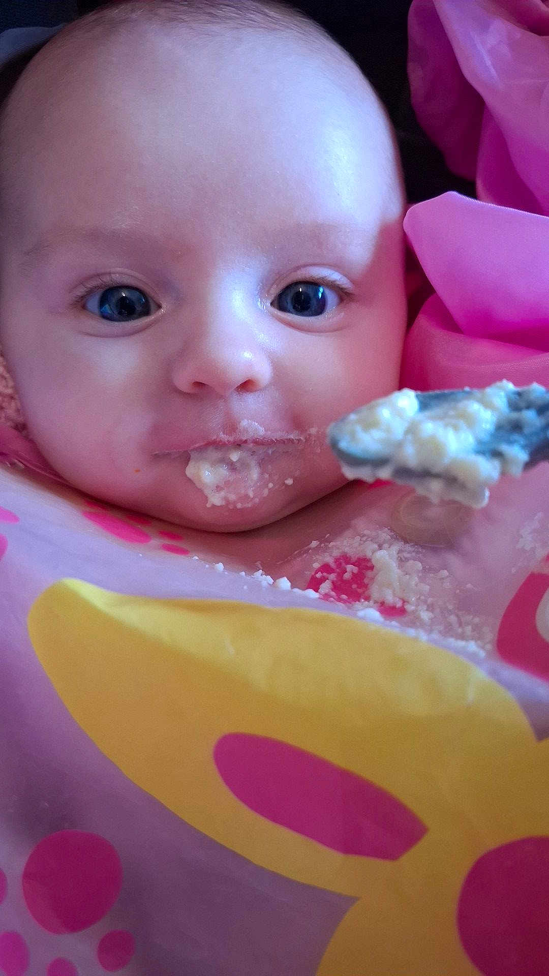 Khloè is registered to the contest to win money with this photo: baby, bib, blue_eyes, child, close_up, cute, expression, face, feeding, food, indoor, infant, messy, person, pink, portrait, skin, spoon, yellow, young