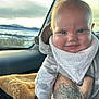 Ellis is registered to the contest to win money with this photo: baby, smiling, infant, hand, tattoo, car_interior, blanket, clothing, window, outdoor_view, face, person, seat, headwear, bib, skin, expression, winter, warm_clothing, transport