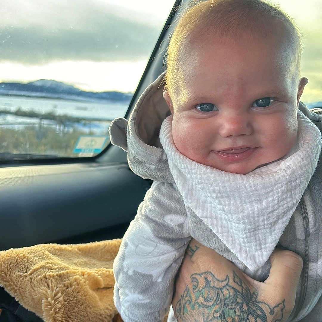 Ellis is registered to the contest to win money with this photo: baby, bib, blanket, car_interior, clothing, expression, face, hand, headwear, infant, outdoor_view, person, seat, skin, smiling, tattoo, transport, warm_clothing, window, winter