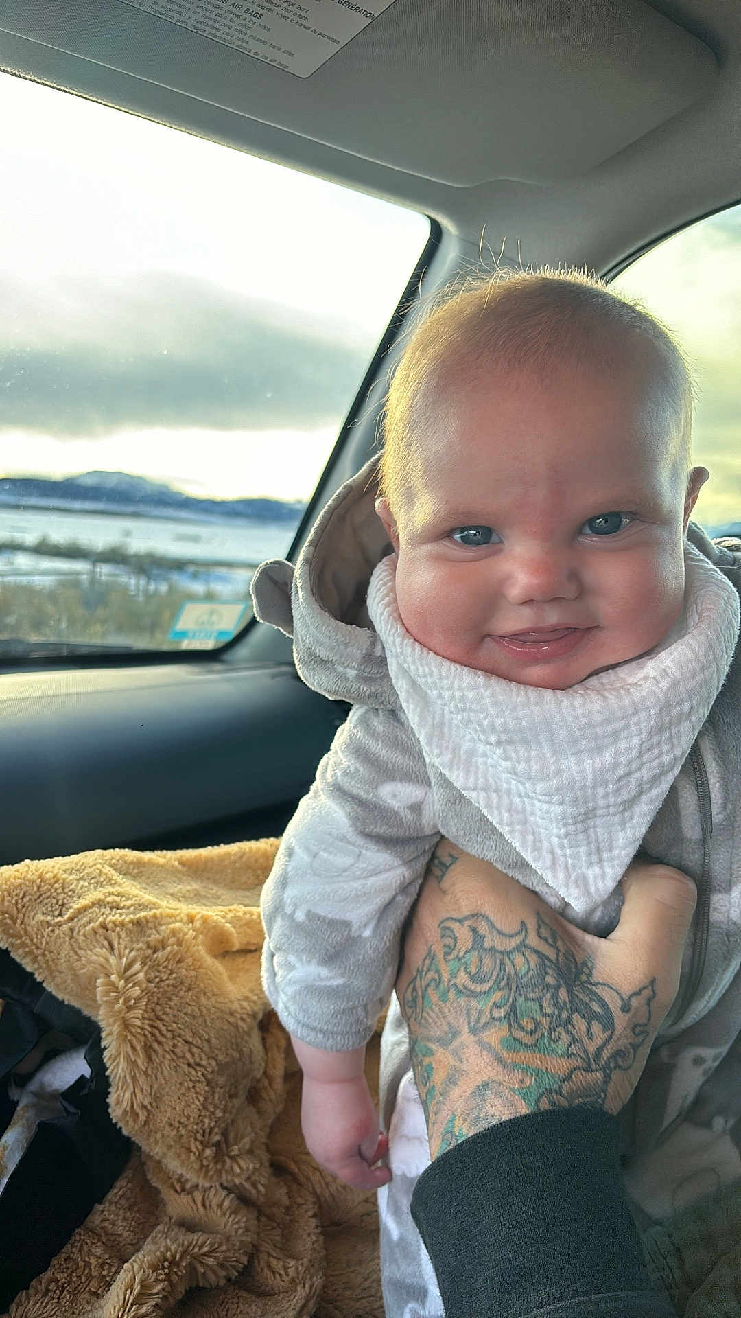 Ellis is registered to the contest to win money with this photo: baby, smiling, infant, hand, tattoo, car_interior, blanket, clothing, window, outdoor_view, face, person, seat, headwear, bib, skin, expression, winter, warm_clothing, transport
