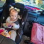 toddler, car_seat, juice_box, plush_toy, car_interior, stroller, yellow_bows, child, seatbelt, window, trees, sunlight, road, clothing, hair, face, vehicle, seat, toy, pink