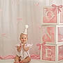 baby, party_hat, clapping, pink, white, birthday, balloons, blocks, bows, smiling, child, sitting, celebration, cute, one_year, decorations, indoors, soft_lighting, portrait, happy