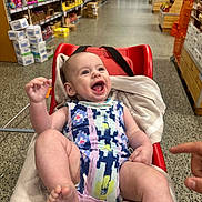 Xavier is registered to the contest to win money with this photo: baby, shopping_cart, grocery_store, aisle, smiling, happy, infant, towel, clothing, red, person, shopping, retail, floor, shelves, products, indoor, foot, hand, face