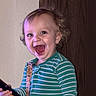 child, curly_hair, door, doorknob, eyes, hand, happy, indoor, laughing, open_mouth, pacifier, pacifier_clip, pajamas, portrait, remote_control, smiling, standing, striped_pajamas, toddler, wall