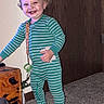 baby_sock, carpet, child, curly_hair, doorknob, green_pajamas, happy, household, indoor, nightstand, onesie, pacifier_clip, pajamas, smiling, standing, striped_clothing, teeth, toddler, toy, wooden_door