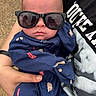 Baylor is registered to the contest to win money with this photo: baby, sunglasses, blue_onesie, baseball_theme, hand, adult, person, reflection, outdoor, clothing, portrait, infant, cool, fashion, babywear, casual, holding, human, face, closeup