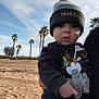 baby, child, hat, pacifier, toy, beach, sand, palm_trees, outdoor, sky, clouds, person, clothing, hand, face, expression, warm_clothing, nature, daylight, holding