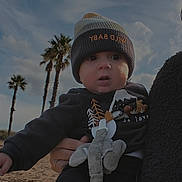 Elio a rejoint le concours — aidez-le/la à gagner de superbes lots ! baby, child, infant, beanie, pacifier, outdoor, beach, sand, cloudy_sky, palm_trees, clothing, cute, person, holding, toy, winter_clothes, face, expression, curious, nature