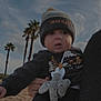 baby, child, infant, beanie, pacifier, outdoor, beach, sand, cloudy_sky, palm_trees, clothing, cute, person, holding, toy, winter_clothes, face, expression, curious, nature