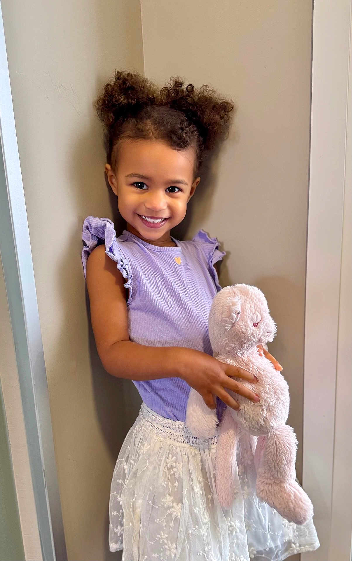Ciara participe au concours pour gagner de l'argent avec cette photo : child, girl, smile, curly_hair, pigtails, lavender_top, white_skirt, plush_bunny, stuffed_animal, holding_toy, portrait, indoor, happy, cute, young_child, eyes, face, person, wall, fashion