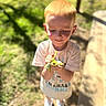 child, boy, red_hair, outdoor, sunlight, greenery, plant, nature, curious, smile, short_sleeve_shirt, white_shoes, daylight, grass, shadow, young, standing, holding, casual_clothing, happy