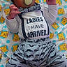 newborn, baby, infant, child, portrait, lying_down, pacifier, smiling, face, eyes, hair, onesie, shirt, mustache_pattern, blanket, teddy_bear_pattern, bedroom, indoor, text_on_clothing, safe