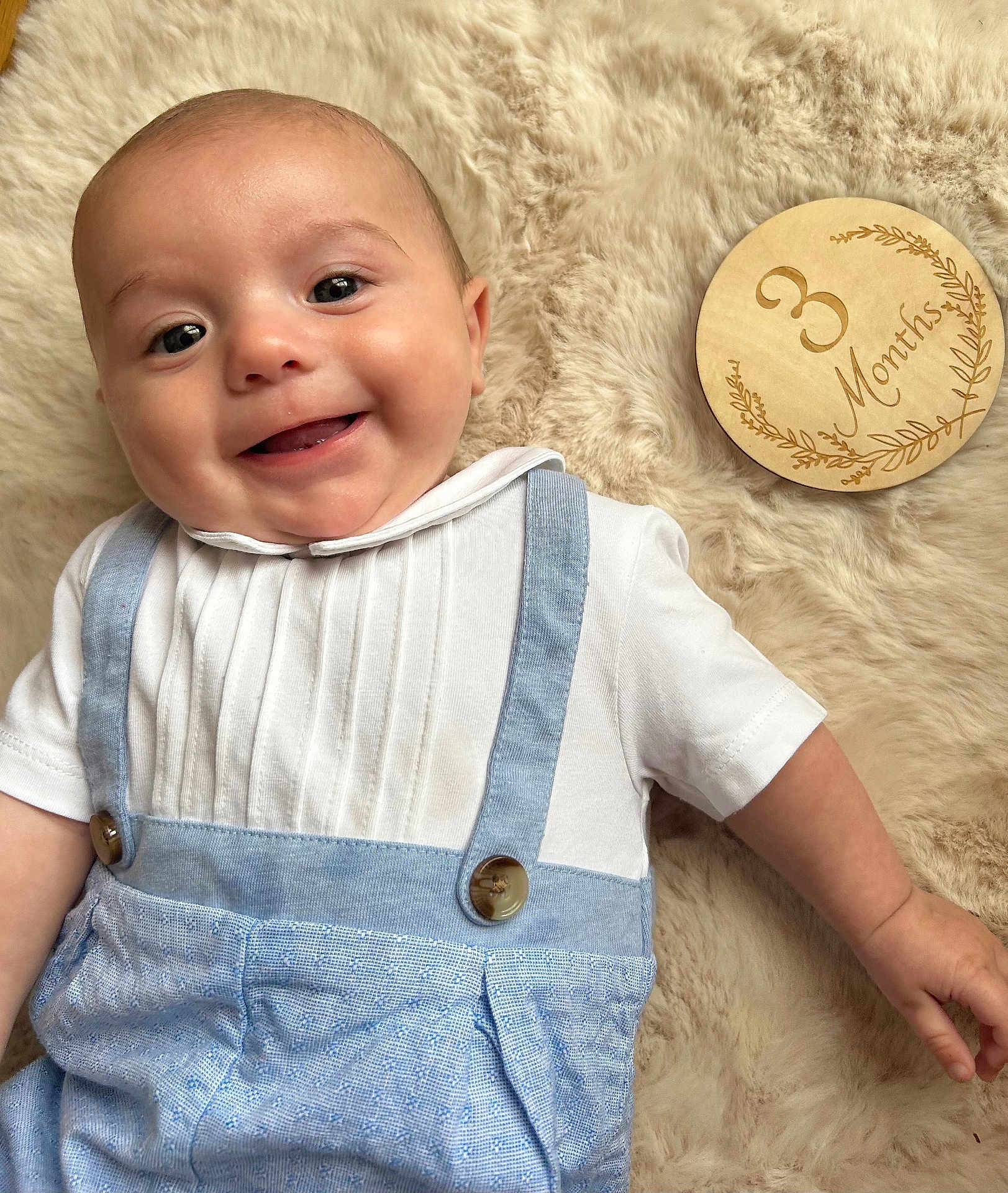 Gabriel participe au concours pour gagner de l'argent avec cette photo : baby, infant, smile, face, clothing, suspenders, white_shirt, blue_pants, wooden_plaque, 3_months, milestone, blanket, fur_texture, cute, portrait, lying_down, happy, child, skin, hand