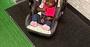 Jazmine is registered to the contest to win money with this photo: baby, car_seat, pink_headband, pink_bib, indoor, green_carpet, black_mat, white_cabinet, brick_wall, plant, child, footwear, clothing, seatbelt, flooring, person, infant, cute, toddler, relaxing