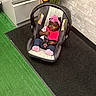 baby, car_seat, pink_headband, pink_bib, indoor, green_carpet, black_mat, white_cabinet, brick_wall, plant, child, footwear, clothing, seatbelt, flooring, person, infant, cute, toddler, relaxing
