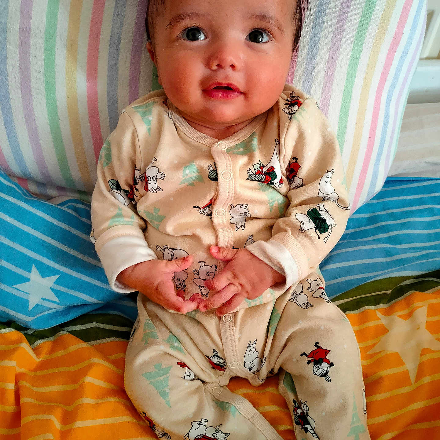 Rayyan is registered to the contest to win money with this photo: baby, bed, child, clothing, cozy, cute, eyes, face, hair, hands, indoors, infant, pajamas, portrait, resting, sitting, smiling, striped_blanket, striped_pillow, young_child