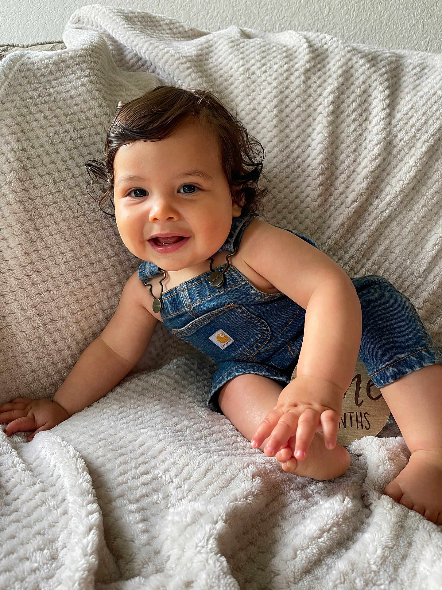 Crew is registered to the contest to win money with this photo: baby, child, denim_overalls, smiling, curly_hair, blanket, texture, indoors, cute, happy, person, infant, skin, hand, foot, portrait, sitting, cozy, comfortable, clothing