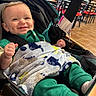 Hendrix is registered to the contest to win money with this photo: baby, smile, stroller, blanket, green_clothing, indoor, dining_area, chairs, red_chairs, black_chairs, happy, infant, cute, person, face, hands, cozy, warm_lighting, floor_tiles, window