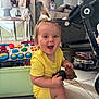 toddler, child, yellow_clothing, toy, smiling, indoor, floor, stroller, living_room, table, lamp, photo_frame, plastic_toys, happy, person, hair_tied, white_shoes, couch, water_bottle, carpet