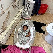 Luna is registered to the contest to win money with this photo: baby, infant, child, bouncer, bathroom, laundry_basket, clothing, towel, toilet, tile_floor, bathroom_mat, pink_blanket, gray_outfit, hood, sitting, portrait, face, indoor, household, resting