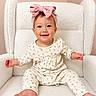 baby, child, smiling, pink_bow, headband, white_chair, fluffy_chair, cute, happy, sitting, infant, long_sleeve_outfit, patterned_clothing, barefoot, indoor, portrait, young_child, light_background, cozy, playful