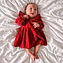 baby, infant, newborn, child, red_dress, dress, bed, white_bed, blanket, lying_down, portrait, smiling, hands, feet, cozy, fabric_texture, adorable, indoor, soft_lighting, bedding